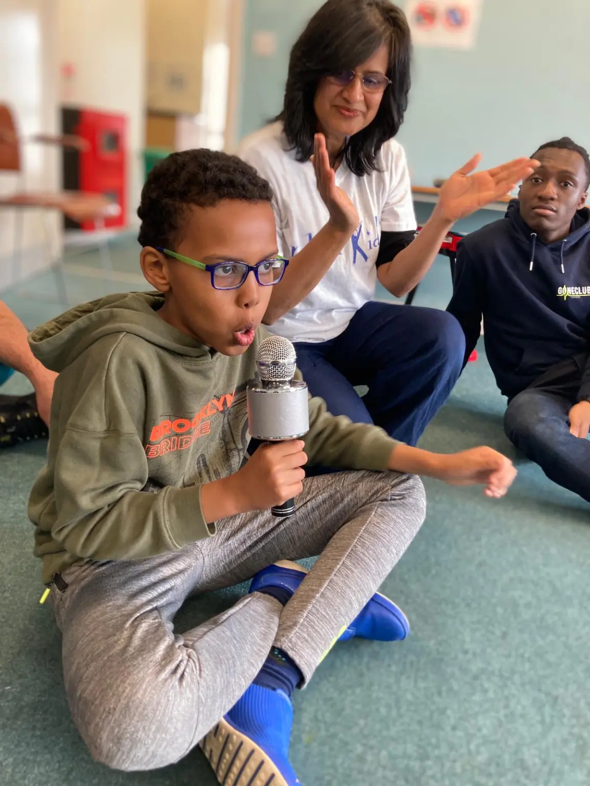 A group of people are sat on the floor, one boy is singing into a microphone whilst the others clap