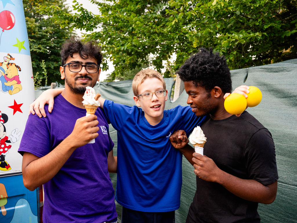 Three older boys are standing together smiling and chatting. The boy in the middle has his arms around the other two, they are eating ice creams.