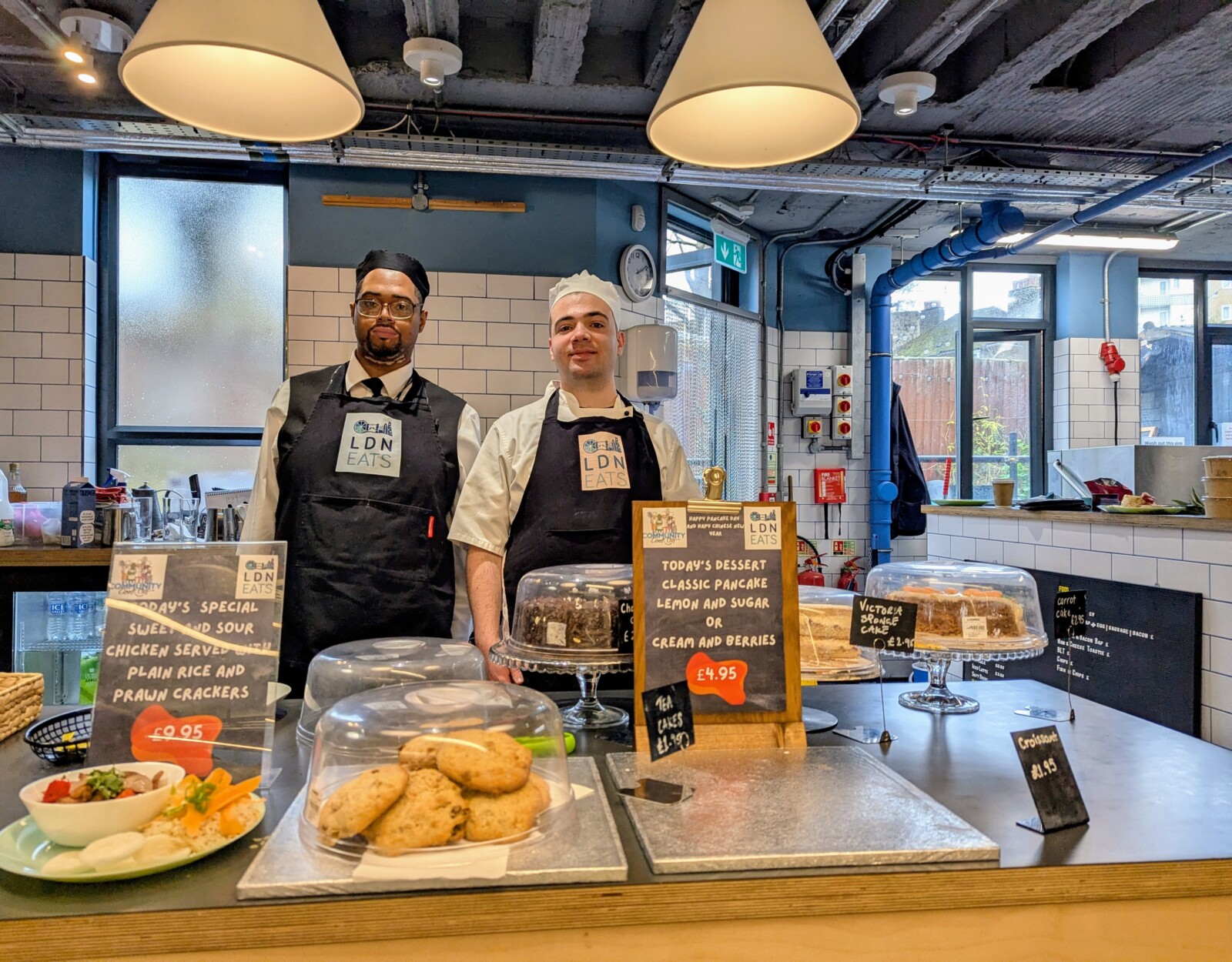 Two people in chefs' aprons and hats behind a display of cakes and pastries